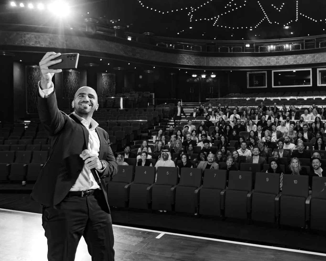 Ahmed El-Gamal on a theater stage, holding up a phone to take a picture with a seated audience of colleagues behind him