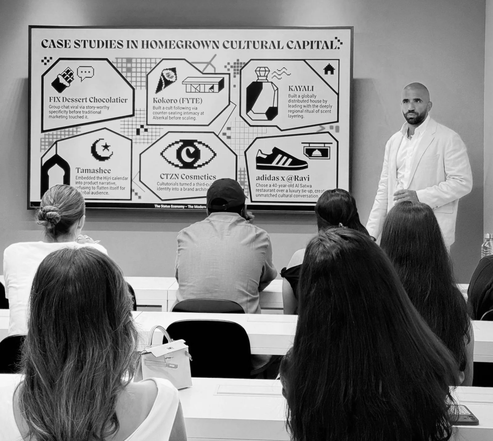 Ahmed El-Gamal in a white suit teaching a class, beside a screen titled Case studies in homegrown cultural capital