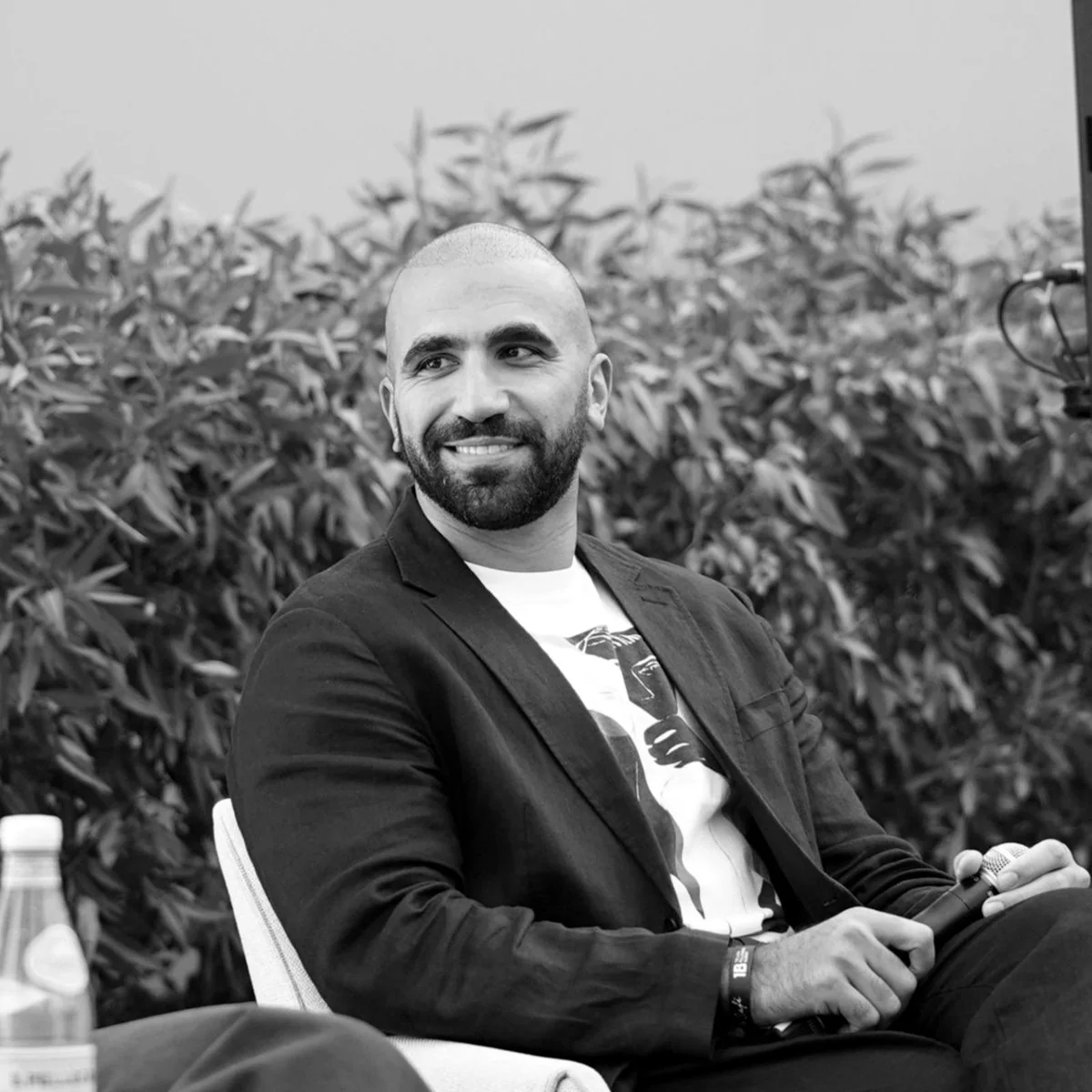 Ahmed El-Gamal seated on an outdoor stage, smiling, holding a microphone